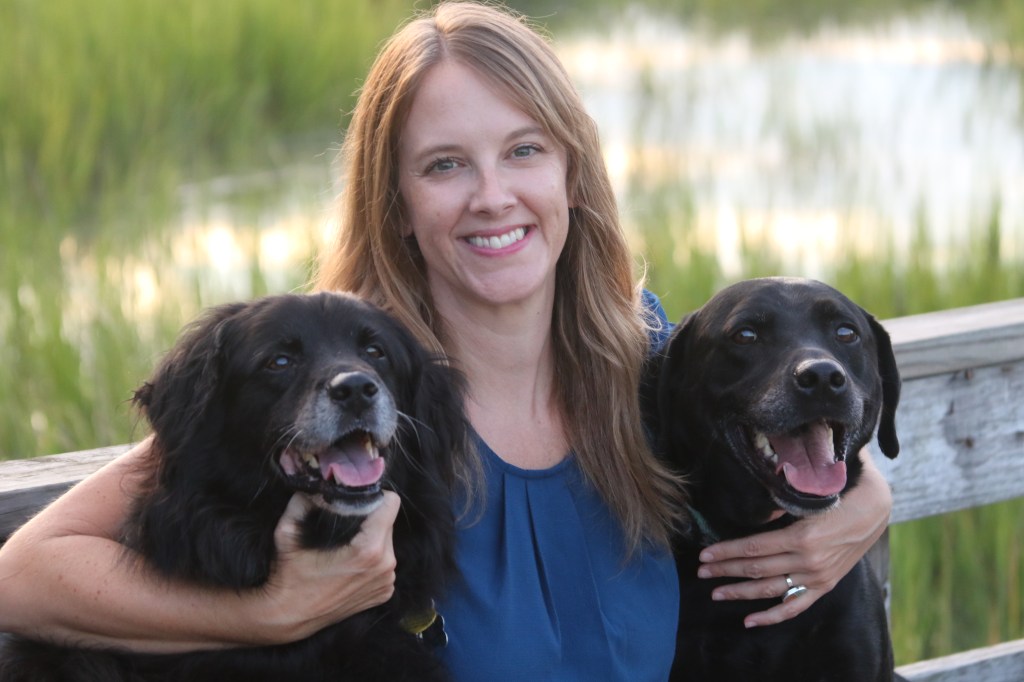 Julie sits on a bench in front of a marsh smiling, with her arms wrapped around her two black labradors.